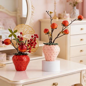 May include: Two ceramic strawberry-shaped vases, one red and one pink, each holding decorative branches with fruit. The red vase has a gold fan-shaped accent. The pink vase sits on a white pedestal, and both are on a cream-colored surface.