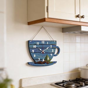 May include: A blue wooden wall clock shaped like a coffee cup, with white numerals and hands. The clock features coffee beans and leaves on the saucer. The clock is hanging on a white wall in a kitchen.