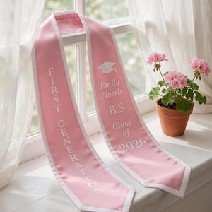 May include: A pink graduation sash with white edging and text. The sash says "FIRST GENERATION" on one side and "Emily Norris B.S. Class of 2026" with a mortarboard graphic on the other. A small potted plant with pink flowers is also visible.