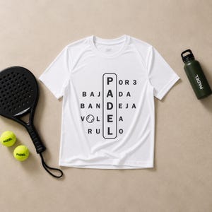 May include: A white short-sleeved t-shirt featuring a black graphic with the word PADEL vertically centred, surrounded by the words POR 3, BAJADA, BANDEJA, VOLEA, and RULO, accompanied by a black padel racket, two yellow padel balls, and a dark green bottle.