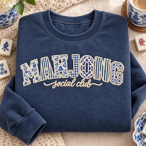 Puede incluir: Una sudadera azul marino de cuello redondo con el texto bordado MAHJONG en letras grandes rellenas de varios patrones azules y blancos, y las palabras social club en una fuente cursiva blanca más pequeña centrada debajo.