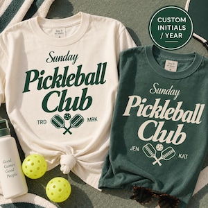 Puede incluir: Una camiseta color crema con texto verde oscuro que dice Sunday Pickleball Club y un gráfico de paleta, y una camiseta verde oscuro con texto crema que dice Sunday Pickleball Club, un gráfico de paleta y los nombres JEN y KAT, más dos pelotas amarillas.