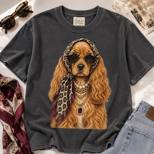 Puede incluir: Una camiseta de cuello redondo gris carbón con un gráfico de un perro spaniel dorado que lleva un pañuelo con estampado de leopardo, gafas de sol oscuras y collares dorados superpuestos con perlas y un colgante con la letra C.