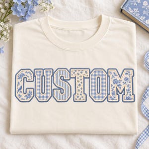 Puede incluir: Una camiseta de cuello redondo color crema con la palabra CUSTOM en letras grandes de bloque. Cada letra está rellena con un patrón azul y blanco diferente, incluyendo estampados florales, cuadros vichy y rayas verticales, delineado con hilo azul.