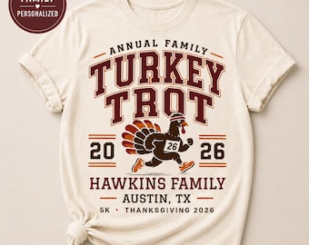 May include: A cream-colored t-shirt featuring a graphic with the text ANNUAL FAMILY TURKEY TROT 20 26 HAWKINS FAMILY AUSTIN, TX 5K THANKSGIVING 2026. The design includes a running turkey wearing a headband and a race bib with the number 26.