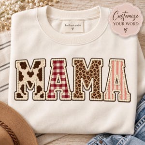 Puede incluir: Una sudadera de cuello redondo color crema con la palabra MAMA en letras grandes. Cada letra tiene un patrón único: manchas de vaca, cuadros vichy rojos y blancos, estampado de leopardo y rayas verticales rosas y blancas, con bordes cosidos.
