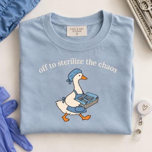 Op de afbeelding: Een lichtblauw t-shirt met een afbeelding van een witte gans die een blauwe operatiemuts en schort draagt en een mandje met medische instrumenten vasthoudt. De tekst boven de gans luidt "off to sterilize the chaos". Het label toont "LINE B ART STUDIO".