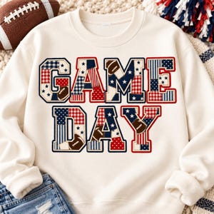 Puede incluir: Una sudadera de cuello redondo color crema con el texto GAME DAY en letras grandes de bloque. Las letras están rellenas con un patrón de retazos en colores rojo, blanco y azul, incluyendo estrellas, rayas, lunares y pequeños gráficos de balones.