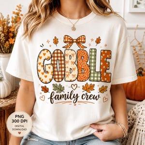 Puede incluir: Una camiseta blanca de manga corta con el texto GOBBLE en letras grandes con varios patrones, rematadas con un lazo a cuadros. Debajo, el texto family crew está escrito en cursiva, rodeado de pequeñas ilustraciones de corazones y hojas.