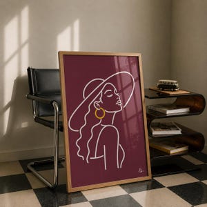 Puede incluir: Una lámina de arte enmarcada que presenta un dibujo de líneas blancas de una persona con un sombrero de ala ancha y un pendiente de aro grande sobre un fondo burdeos intenso. La obra incluye una pequeña firma en la esquina inferior derecha.