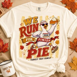 May include: An off-white t-shirt featuring a graphic of a white goose wearing sunglasses and a headband, running while holding a slice of pumpkin pie. The text reads WE RUN FOR FINISH PIE 2026 TURKEY TROT FAMILY. The design includes red and orange accents.