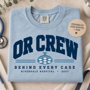 Puede incluir: Una camiseta de cuello redondo azul claro con texto azul oscuro que dice OR CREW BEHIND EVERY CASE RIVERDALE HOSPITAL 2027. Debajo del texto OR CREW hay un gráfico de luz quirúrgica azul claro. La etiqueta del cuello muestra Comfort Colors.