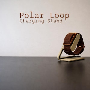 Könnte beinhalten: Ein brauner Polar Loop Ladeständer mit einer Uhr. Die Uhr hat ein braunes Armband und ein beiges Innenteil. Der Ständer ist beige und hat ein minimalistisches Design. Der Text "Polar Loop Charging Stand" ist braun.