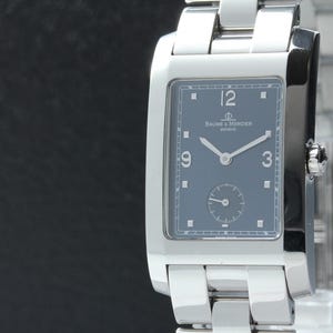 May include: A rectangular stainless steel wristwatch with a polished silver-tone finish and a dark blue dial. The watch face features silver-tone hands, Arabic numerals at 3, 9, and 12 o'clock, a small seconds sub-dial, and the Baume & Mercier logo.