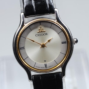May include: A Credor wristwatch featuring a round silver dial with gold-tone hands and indices, encased in a silver-tone metal frame with a gold-tone inner bezel. The watch includes a black leather strap with a textured finish and a crown on the side.