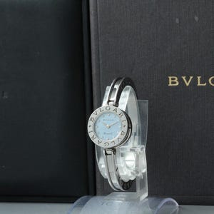 May include: A silver-toned Bvlgari wristwatch featuring a round case with a light blue dial and the brand name engraved on the bezel. The watch has a slim, open-link metal bracelet and is displayed in front of a black branded presentation box.