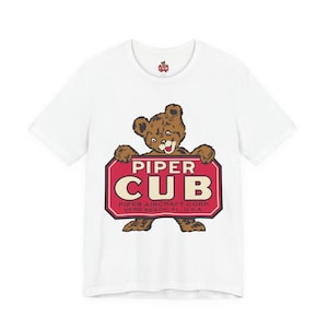 May include: White t-shirt with a brown bear holding a red sign that reads "PIPER CUB" in white. The bear has a smiling expression. The text "PIPER AIRCRAFT CORP. VERO BEACH, FL. U.S.A." is below the sign.