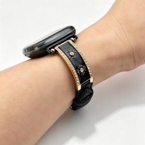 May include: A black smartwatch featuring a decorative metal band segment with a glossy black center inlay, gold-tone edges, and small clear crystals. The band segment displays two gold-tone sunburst or floral motifs and connects to a black strap.