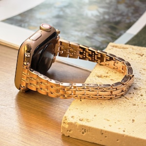 Op de afbeelding: Een roségouden smartwatch met een bijpassende metalen band. De band is gemaakt van kleine, rechthoekige schakels. Het horloge ligt op een lichtgekleurd, getextureerd oppervlak, met een vage achtergrond.