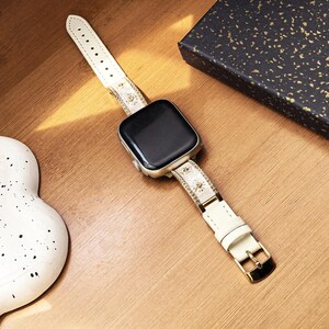 K&ouml;nnte beinhalten: Eine Smartwatch mit einem rechteckigen schwarzen Bildschirm und einem silberfarbenen Geh&auml;use. Das Armband besteht aus cremefarbenem Material mit kleinen goldenen Sonnen- und Augenmotiven, akzentuiert durch d&uuml;nne goldene R&auml;nder und eine goldene Schlie&szlig;e.