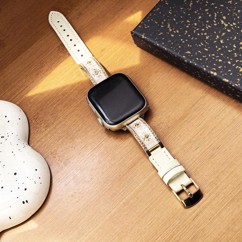 K&ouml;nnte beinhalten: Eine Smartwatch mit einem rechteckigen schwarzen Bildschirm und einem silberfarbenen Geh&auml;use. Das Armband besteht aus cremefarbenem Material mit kleinen goldenen Sonnen- und Augenmotiven, akzentuiert durch d&uuml;nne goldene R&auml;nder und eine goldene Schlie&szlig;e.