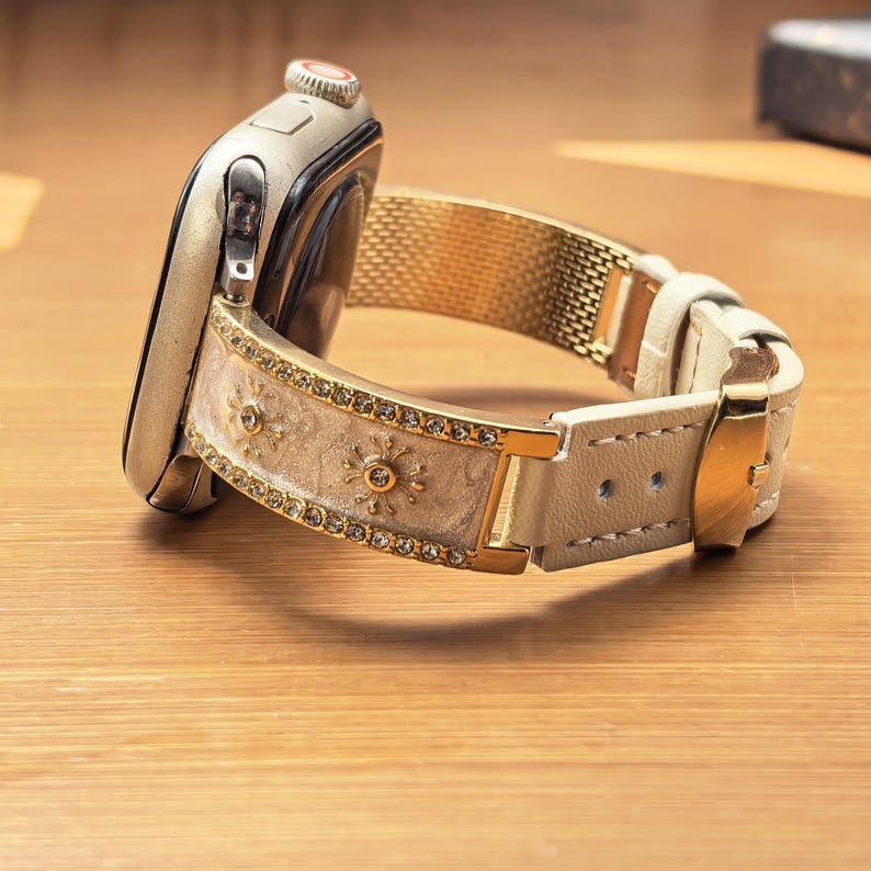 K&ouml;nnte beinhalten: Eine Smartwatch mit Metallgeh&auml;use und dekorativem Armband. Das Armband hat ein cremefarbenes Mittelteil mit goldenen Sonnenmotiven, das von Reihen klarer Kristalle ges&auml;umt ist, und geht in ein glattes, cremefarbenes Lederarmband mit Goldschnalle &uuml;ber.