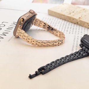 May include: A rose gold metal watch band attached to a smartwatch case, featuring a multi-link design with a polished finish. A separate black metal watch band with a similar multi-link construction is positioned in the foreground.