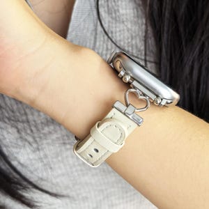 Op de afbeelding: Een zilveren smartwatch met een crèmekleurige leren band. De band heeft een hartvormige connector en een gespsluiting. Het horloge wordt om een pols getoond, waarbij het ontwerp en de stijl van de draagbare technologie worden benadrukt.