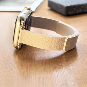 Op de afbeelding: Een goudkleurige smartwatch met een crèmekleurige leren band. De band heeft een textuur en is gebogen, waardoor de wijzerplaat en de banddetails zichtbaar zijn. Het horloge ligt op een houten oppervlak.