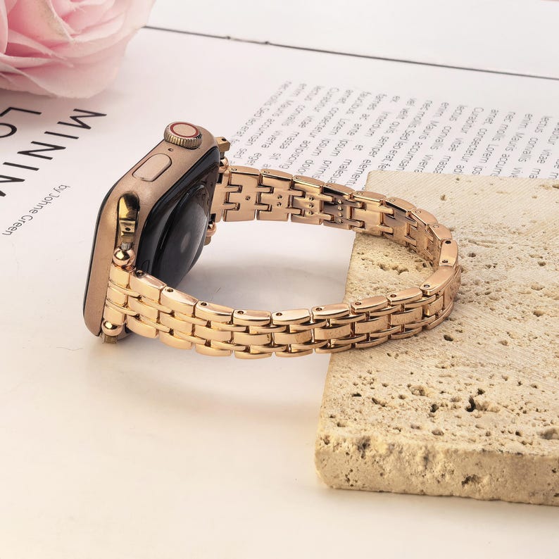 May include: A rose gold smartwatch with a matching metallic link band. The watch features a rectangular case with a side button and a digital crown. The band consists of small, polished rectangular links connected in a multi-row pattern.