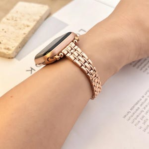 May include: A rose gold metal watch band with a multi-link design attached to a smartwatch case. The band features a polished finish and a secure clasp mechanism, providing a sleek look that wraps around the wrist with a flexible, articulated structure.