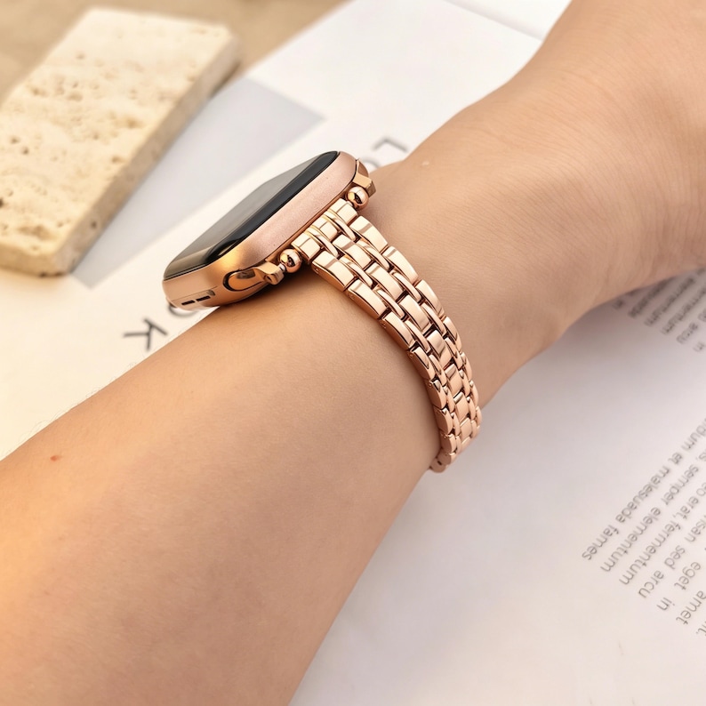 May include: A rose gold metal watch band with a multi-link design attached to a smartwatch case. The band features a polished finish and a secure clasp mechanism, providing a sleek look that wraps around the wrist with a flexible, articulated structure.
