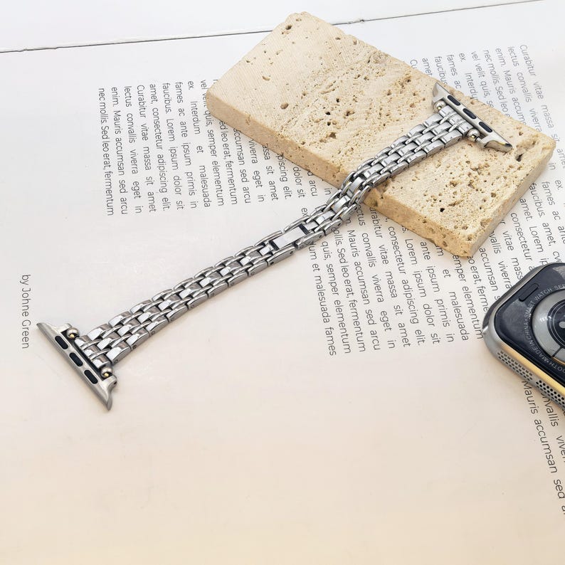 May include: A silver-toned metal link watch band featuring a multi-row chain design with integrated connectors at both ends. The band is partially resting on a rectangular, porous, beige stone block next to a partial view of a smartwatch.
