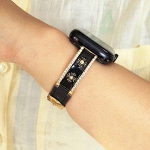 K&ouml;nnte beinhalten: Ein schwarzes Smartwatch-Armband mit einem mittigen, gl&auml;nzend schwarzen Einsatz, der mit zwei goldenen Sonnenmotiven verziert ist. Das Armband ist von zwei Reihen kleiner, klarer, funkelnder Kristalle in einer goldenen Fassung umrandet.