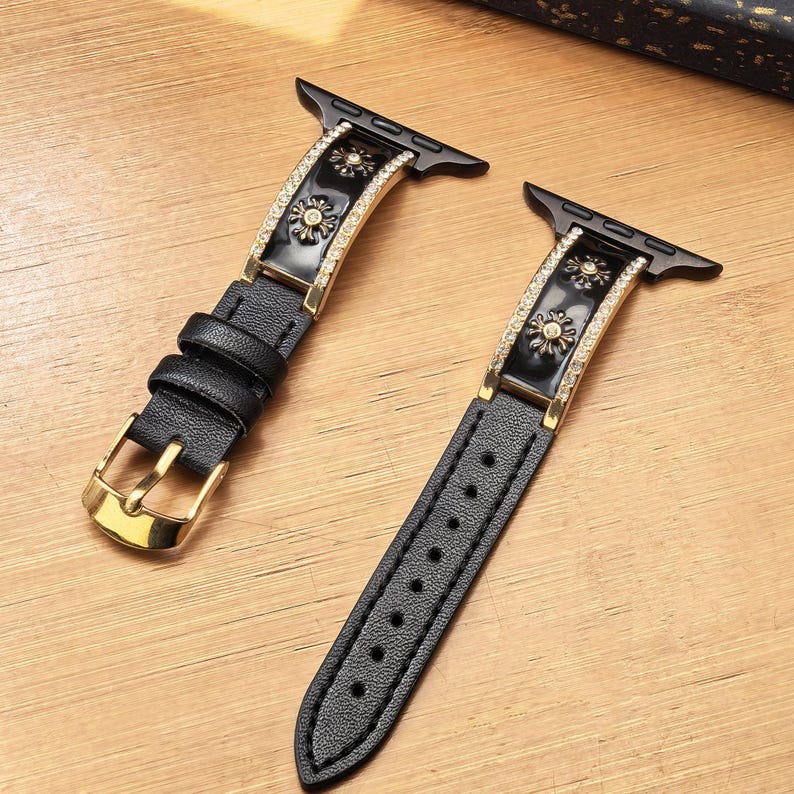 K&ouml;nnte beinhalten: Ein schwarzes Lederuhrenarmband mit goldfarbener Schlie&szlig;e und Verbindungsst&uuml;cken. Das Band verf&uuml;gt &uuml;ber eine mittige Einlage aus schwarzem Emaille mit goldfarbenen Blumenmotiven, die beidseitig von Reihen kleiner klarer Kristalle ges&auml;umt ist.