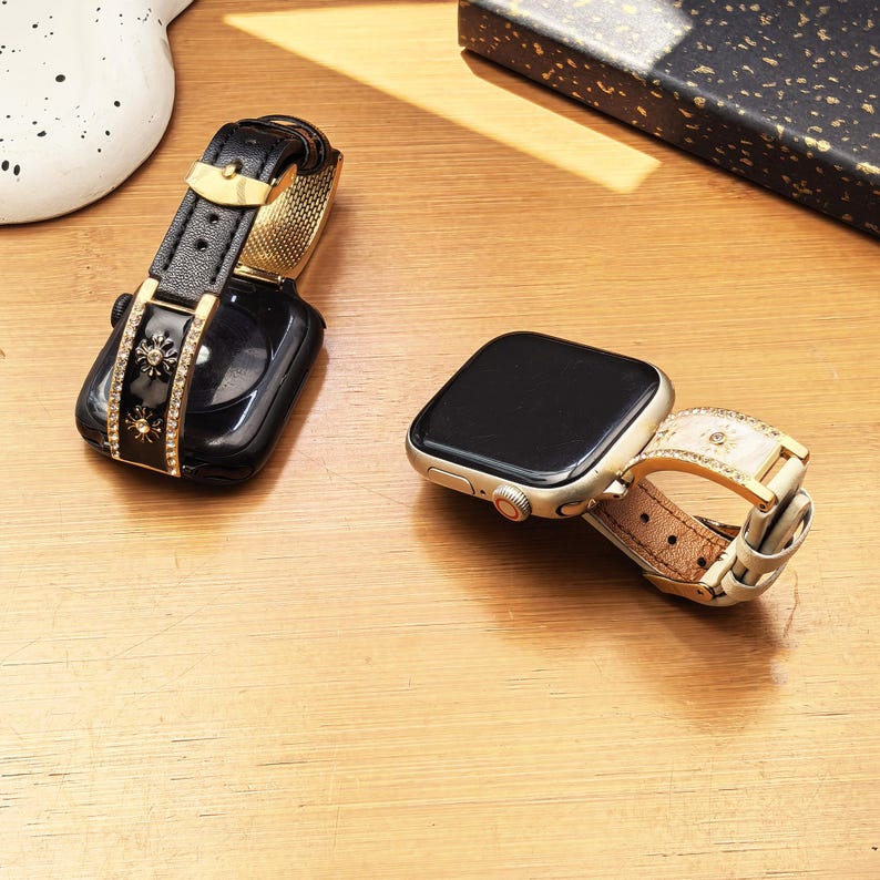 K&ouml;nnte beinhalten: Zwei Smartwatches mit dekorativen Armb&auml;ndern. Eine Uhr hat ein schwarzes Armband mit goldenen Sonnenmotiven und klaren Kristallen. Die andere Uhr hat ein cremefarbenes Armband mit &auml;hnlichen goldenen Sonnenakzenten und Kristallen sowie einem Goldfutter.