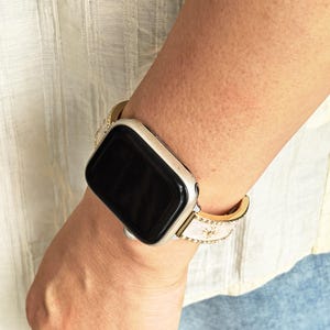 K&ouml;nnte beinhalten: Eine Smartwatch mit einem rechteckigen schwarzen Bildschirm und einem silbernen Metallgeh&auml;use. Das Armband ist wei&szlig; mit goldfarbenen Kanten und weist ein wiederkehrendes Muster aus kleinen, runden goldfarbenen Nieten entlang des Bandes auf.