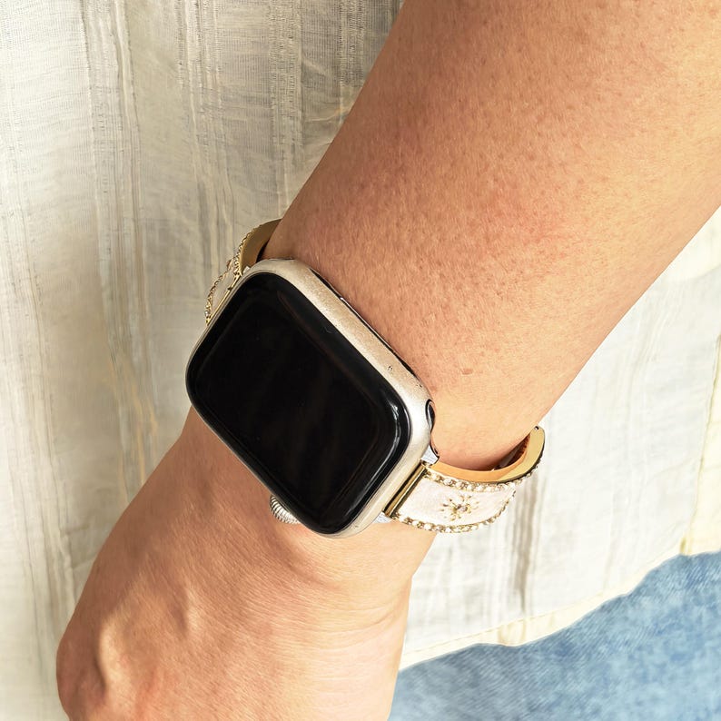 K&ouml;nnte beinhalten: Eine Smartwatch mit einem rechteckigen schwarzen Bildschirm und einem silbernen Metallgeh&auml;use. Das Armband ist wei&szlig; mit goldfarbenen Kanten und weist ein wiederkehrendes Muster aus kleinen, runden goldfarbenen Nieten entlang des Bandes auf.
