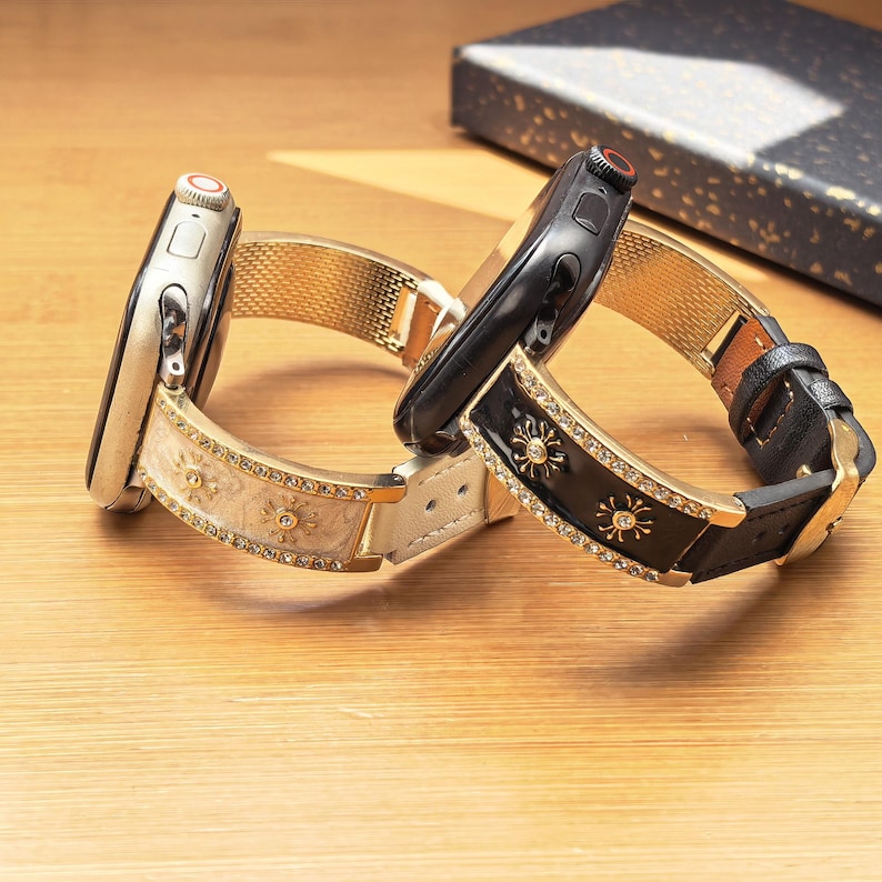 K&ouml;nnte beinhalten: Zwei Smartwatches mit dekorativen Armb&auml;ndern. Ein Armband ist cremefarben mit goldenen Sonnenmotiven und klaren Kristallen. Das andere Armband ist schwarz mit goldenen Sonnenmotiven, klaren Kristallen, einem Lederriemen und einer Schlie&szlig;e.
