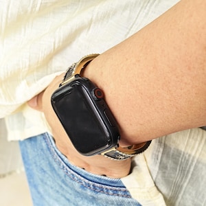 K&ouml;nnte beinhalten: Eine schwarze Smartwatch mit rechteckigem Display und roter digitaler Krone, ausgestattet mit einem goldfarbenen Metallarmband, das an den R&auml;ndern mit kleinen, klaren, funkelnden Kristallen besetzt ist, getragen am Handgelenk mit blauem Jeansstoff.