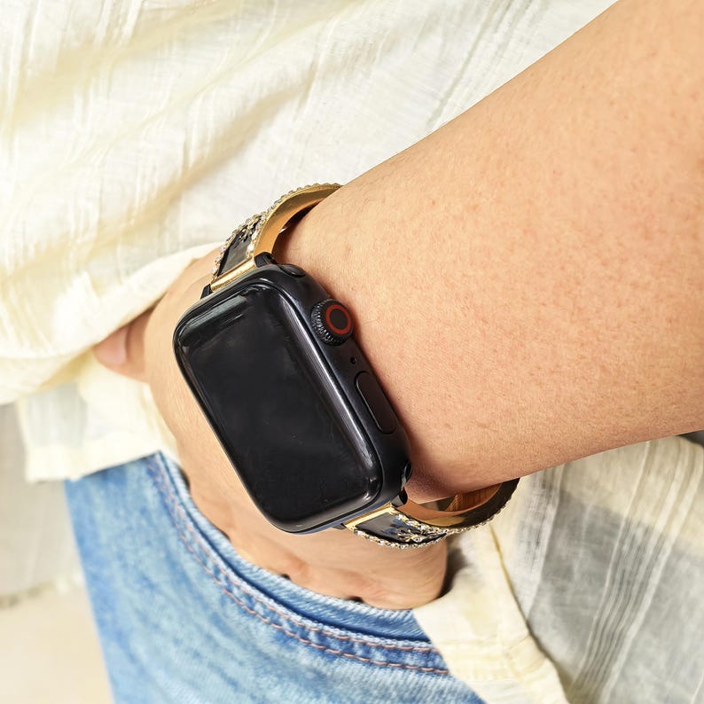 K&ouml;nnte beinhalten: Eine schwarze Smartwatch mit rechteckigem Display und roter digitaler Krone, ausgestattet mit einem goldfarbenen Metallarmband, das an den R&auml;ndern mit kleinen, klaren, funkelnden Kristallen besetzt ist, getragen am Handgelenk mit blauem Jeansstoff.
