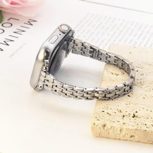 May include: A silver-toned metal watch band attached to a silver smartwatch case. The band features a multi-link design with small, polished rectangular segments connected by rounded links, creating a flexible and structured metallic appearance.