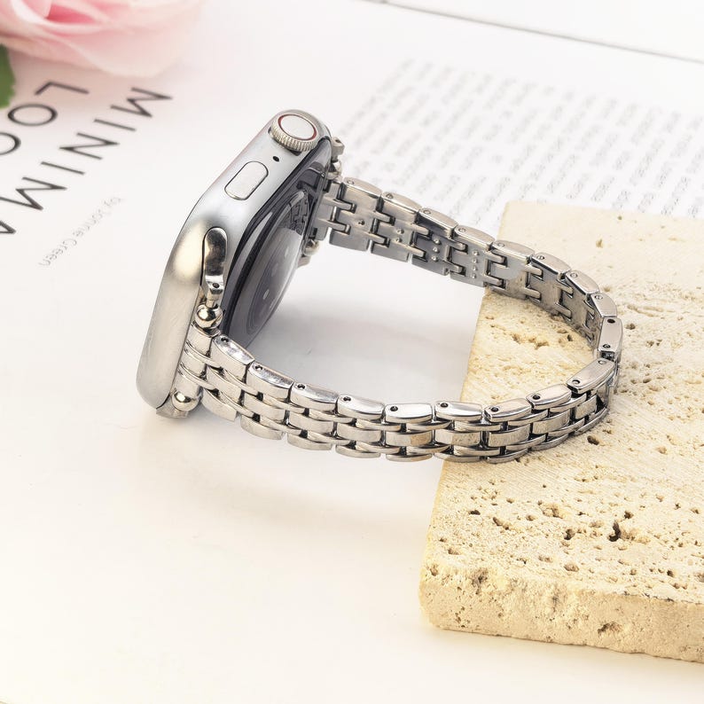 May include: A silver-toned metal watch band attached to a silver smartwatch case. The band features a multi-link design with small, polished rectangular segments connected by rounded links, creating a flexible and structured metallic appearance.
