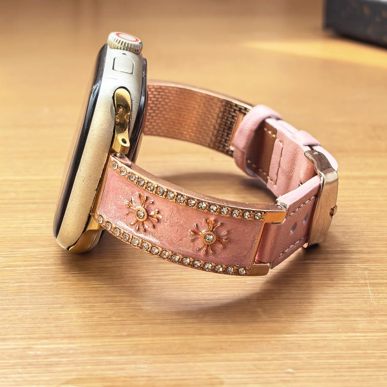K&ouml;nnte beinhalten: Eine Smartwatch mit einem goldfarbenen Metallgeh&auml;use und einem hellrosa Lederarmband. Das Armband ist mit zwei Reihen kleiner, klarer Kristalle an den R&auml;ndern, goldfarbenen sternf&ouml;rmigen Metallakzenten und einer goldfarbenen Schlie&szlig;e versehen.