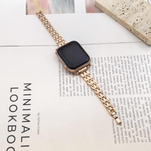 May include: A rose gold metal watch band attached to a rectangular smartwatch with a black screen. The band features a multi-link design with a polished finish, connecting to the watch casing via integrated lugs, creating a cohesive metallic look.