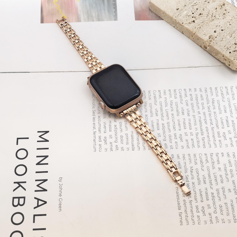 May include: A rose gold metal watch band attached to a rectangular smartwatch with a black screen. The band features a multi-link design with a polished finish, connecting to the watch casing via integrated lugs, creating a cohesive metallic look.