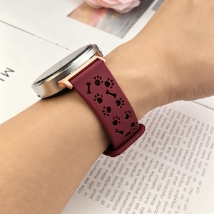 May include: A smartwatch with a metallic silver and rose gold casing featuring a dark maroon silicone band. The band is decorated with a repeating pattern of cut-out paw prints and small bone shapes throughout the length of the flexible material.