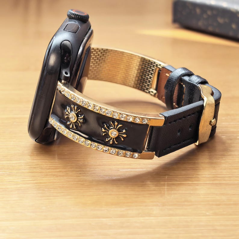 K&ouml;nnte beinhalten: Eine schwarze Smartwatch mit einem dekorativen Armband, das ein schwarzes Emaille-Mittelteil, goldfarbene Metallr&auml;nder mit klaren Kristallen und goldene Sonnenmotive aufweist. Das Band geht in ein schwarzes Lederarmband mit goldener Metallschnalle &uuml;ber.