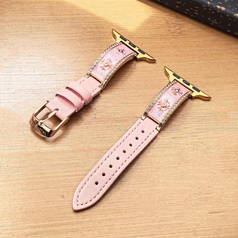 K&ouml;nnte beinhalten: Ein hellrosa Uhrenarmband mit goldfarbenen Metallanschl&uuml;ssen und einer Dornschlie&szlig;e. Das Band verf&uuml;gt &uuml;ber ein Mittelteil mit kleinen, runden, goldfarbenen Blumenmotiven und ist beidseitig mit Reihen klarer, funkelnder Kristalle besetzt.