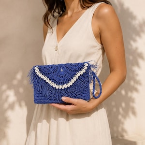 May include: A vibrant royal blue crochet clutch bag featuring a decorative flap adorned with a row of white cowrie shells and fringe detailing. The rectangular bag includes a matching blue braided wrist strap attached to the side for carrying.